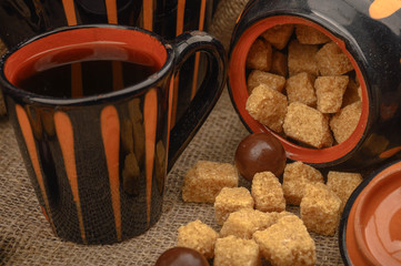 Chunks of brown sugar, a sugar bowl, a mug of tea, and ceramic dishes against a background of coarse homespun cloth. Close up.