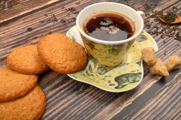 A Cup of black tea, tea leaves, pieces of brown sugar, oatmeal cookies on a wooden background. Close up.