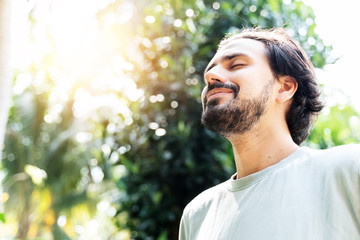 A bearded man is meditating outdoor in the park with face raised up to sky and eyes closed on sunny...
