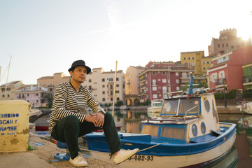 Fototapeta na wymiar Asean young man taking a photo with black hat and smiling on the edge of a city river with boats and views of colorful houses in Valencia, Spain