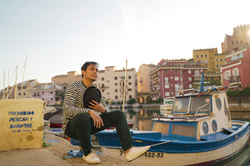 Asean young man taking a photo with black hat and smiling on the edge of a city river with boats and views of colorful houses in Valencia, Spain