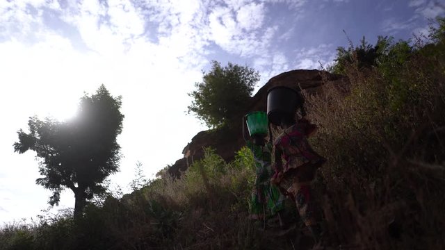 Two Girls With Buckets On Their Head Fighting Their Way Up The Hill Trough Bushes And Scrub