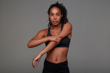Obraz premium Indoor photo of young dark skinned curly lady with casual hairstyle preparing for evening workout after working day, standing against grey background in sporty wear