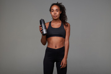 Fototapeta premium Tired pretty young dark skinned curly brunette female with casual hairstyle relaxing after hard training, posing over grey background with black fitness bottle in her arm