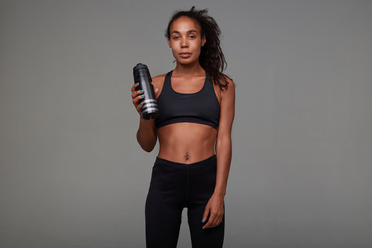 Tired pretty young dark skinned curly brunette female with casual hairstyle relaxing after hard training, posing over grey background with black fitness bottle in her arm © timtimphoto
