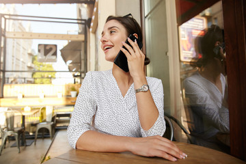 Cheerful young attractive brunette female with bun hairstyle keeping hands on countertop while having pleasant talk on phone, having lunch break in city cafe
