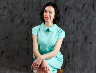 Portrait of a pretty brunette woman in a turquoise dress sitting on a chair on an alternative gray background.