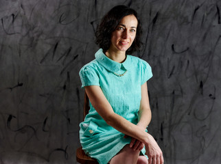 Portrait of a pretty brunette woman in a turquoise dress sitting on a chair on an alternative gray background.