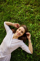 Obraz premium Beautiful young dark haired female in white polka-dot dress lying on green grass and raising hands to her head, being in nice mood and smiling cheerfully at camera