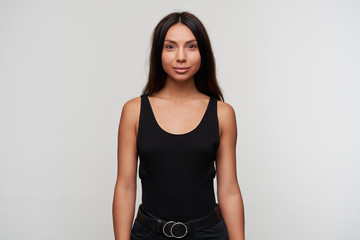 Indoor shot of young lovely dark haired woman with casual hairstyle looking positively at camera and smiling slightly, standing over white background with hands along her body