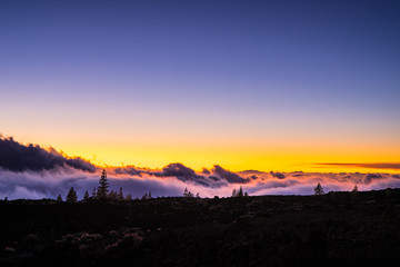 Spain, Tenerife, Orange sky and moonlight shining on arid black lava nature landscape above the clouds in mountains of caldera after sunset by night