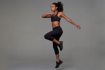 Full-length shot of slim young dark skinned curly brunette female being concentrated while making physical exercises in fitness studio, standing against grey background