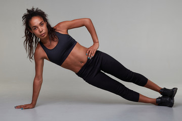 Fototapeta na wymiar Horizontal photo of young sporty dark skinned woman with curly long hair making physical exercises in gym class, wanting to be fit and healthy, isolated over grey background
