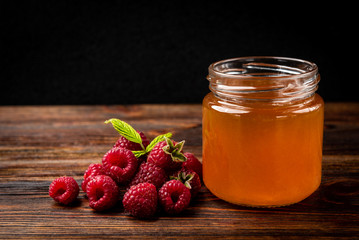  Honey in glass jar and fresh raspberry on dark wooden background.