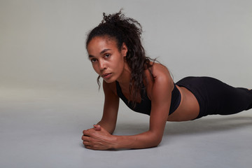 Portrait of young dark haied concentrated curly lady staying in plank while posing over grey background, looking thoughtfully aside with folded lips and keeping arms crossed