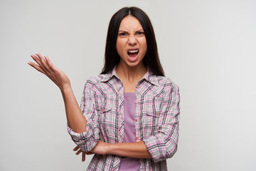 Indignant young dark haired lady with natural makeup raising palm emotionally while standing over white background, looking angily to camera and frowning her face