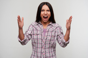 Excited young pretty dark haired female with casual hairstyle looking emotionally to camera with raised hands and keeping mouth wide opened, standing against white background