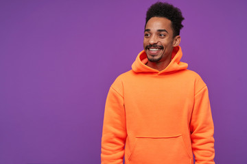 Indoor shot of cheerful young pretty bearded brunette man with dark skin looking aside with charming smile, being in nice mood while posing over purple background