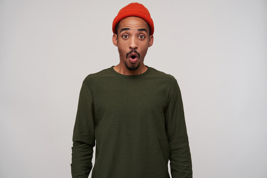 Overwhelmed young attractive bearded brunette male with dark skin looking amazedly at camera with round eyes, wearing red hat and khaki sweater over white background