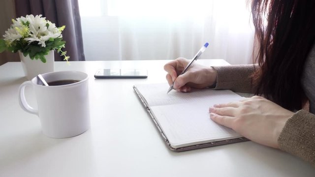 A young woman sits at a table and writes notes in a notebook. A list of important tasks for the week. Close-up