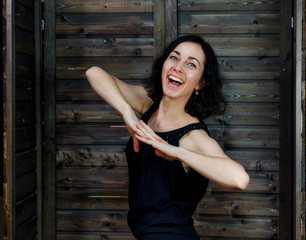 Portrait of a pretty smiling brunette woman in black dress on wooden background in vintage interior. Standing right in front of the camera with vivid emotions.