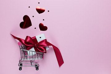Shopping cart with gift box and love hearts on pink background with copy space. Valentine's day.