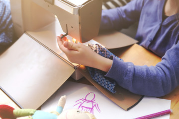Enthusiastic young girl works with a compact sewing machine under supervision of her mother to make a toy dress for her doll - background blanked out blurry