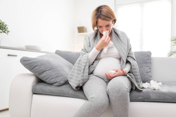 Pregnant woman catching cold, flu, virus, sitting at home on a couch, blowing nose and being sick.