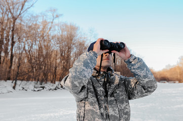 traveler in camouflage clothes looking at winter nature through binoculars