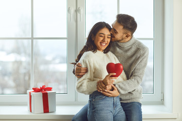 Valentine's day. Beautiful smiling couple gives a heart on a background of a window in a room