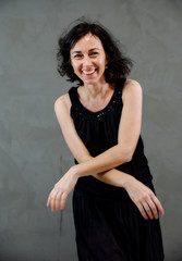 Model Standing right in front of the camera with vivid emotions. Vertical portrait of a cute smiling brunette woman in black dress on gray alternative background.