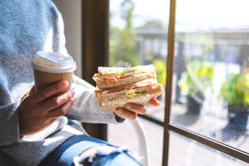 Closeup image of a woman holding and eating whole wheat sandwich and coffee in the morning