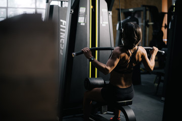 Fototapeta na wymiar Muscular sporty woman using training simulator for pumping back muscles in gym, back of view. Concept of professional training in a modern dark gym.