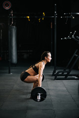 Naklejka na ściany i meble Athletic muscular woman preparing to lift the heavy barbell from the floor, side view. Concept of healthy lifestyle and workouts in a modern dark gym.