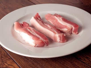 Uncooked raw pork belly slices on a white plate and wooden table.