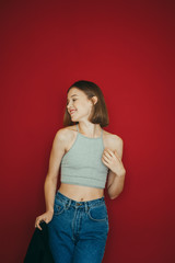 Smiling girl in stylish casual clothes stands on a red background and poses at the camera. Vertical lifestyle photo of positive girl rejoicing, isolated. Happy lady on colored background