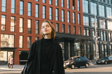 Fototapeta na wymiar Attractive girl in casual fashion poses against the background of modern architecture and the street, looks into the camera with a smile. Young business lady standing on the street of a megapolis