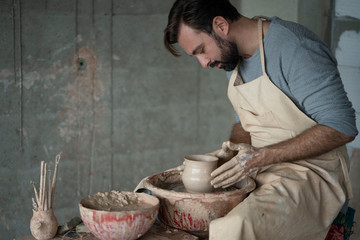 Attractive smiley man with beard making ceramic pot on the pottery wheel. Craftsman creating pottery working on the wheel shaping clay making vase. Close up portrait.