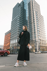 Fototapeta na wymiar Stylish model in black clothes and sneakers stands on street metropolis, looks away and poses for the camera with a serious face.Full length portrait of a fashionable girl on a walk in the city