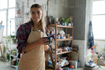 Young lovely smiling girl with blonde hair in apron holding a clay pot in her hands and standing in pottery studio. Pottery workshop. Clay model.
