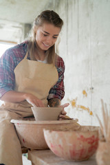 Young beautiful smiley woman in apron holding a clay pot in her hands and siting on bench with pottery wheel in pottery studio . Pottery workshop. Clay model.