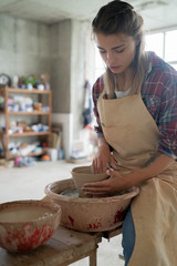 Young beautiful smiley woman in apron holding a clay pot in her hands and siting on bench with pottery wheel in pottery studio . Pottery workshop. Clay model.