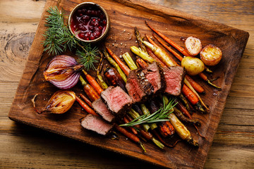 Grilled sliced Venison Steak with baked vegetables and berry sauce on wooden background - obrazy, fototapety, plakaty