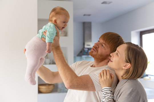 Positive new parents holding and staring at crying baby. Family portrait of couple and cute little child in home interior. Parenthood or tenderness concept
