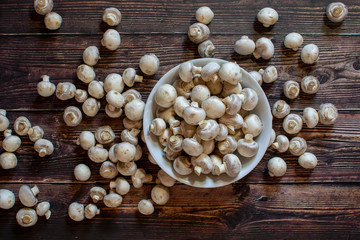 Champignon mushrooms (Agaricus campestris) on a rustic wooden surface, top view. Mushrooms in a white porcelain bowl.