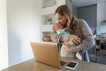 New mother holding and rocking crying baby near laptop. Portrait of young woman and cute little child in home interior. Communication concept