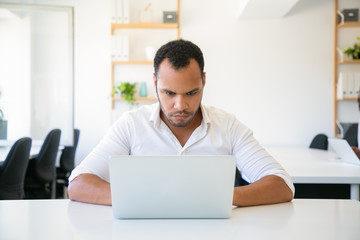 Pensive handsome man using laptop at modern office. Front view of young confident programmer working at office. Technology concept