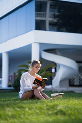Beautiful smart girl with gathered hair sitting on a green lawn and reading book. Girl wearing in bikini and white tunic. The suns rays shining on her.