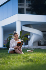 Beautiful smart girl with gathered hair sitting on a green lawn and reading book. Girl wearing in bikini and white tunic. The suns rays shining on her.