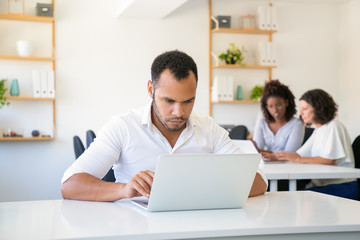 Fototapeta premium Concentrated handsome programmer working with laptop. Front view of confident bearded man using modern device at office. Technology concept